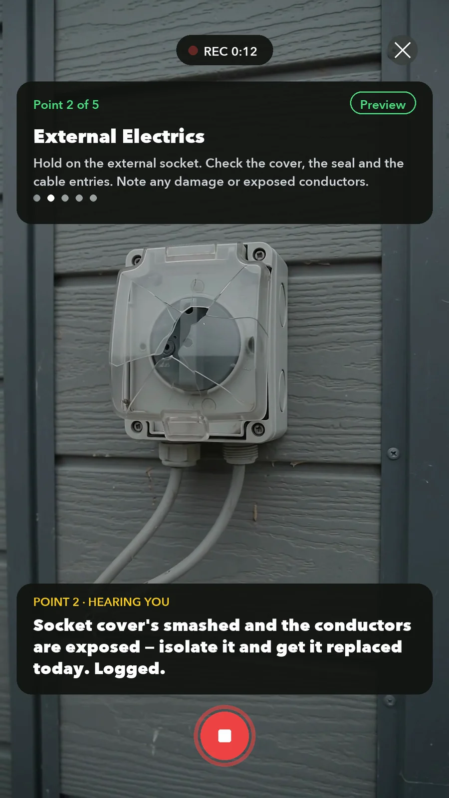 A SiteSurvey Walk Cam inspection on a phone: the engineer films a cracked external socket cover on a building elevation while live voice-to-text writes the observation — 'socket cover's smashed and the conductors are exposed, isolate it and get it replaced today'
